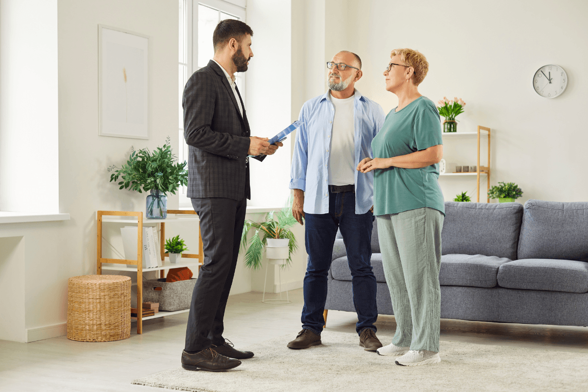 Male talks through home features with a mature senior couple during house visit, focusing on layout and livingroom appeal details.